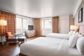 A modern hotel room features two beds, large windows with colorful curtains, a cozy chair, and a TV, creating a welcoming atmosphere.