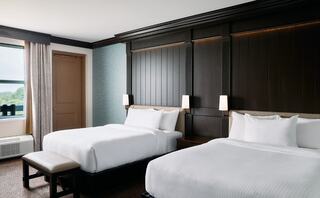 A modern hotel room features two cozy beds, warm wood accents, soft lighting, and a window with a view, creating a welcoming atmosphere.