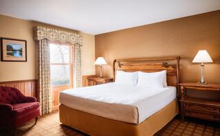 A cozy hotel room features a queen-sized bed, wooden furniture, a vintage armchair, and a window with drapes overlooking a serene view.