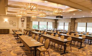 A spacious meeting room features rows of tables and chairs, stylish lighting, and large windows, providing a bright and inviting atmosphere.