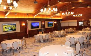 A spacious, elegantly designed banquet room with round tables, decorative chandeliers, and large screens displaying images on the walls.