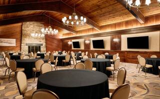 A spacious, elegantly designed banquet room with circular tables, beige chairs, and rustic wood accents, featuring chandeliers and two televisions.