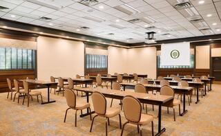 A spacious meeting room with rows of tables and chairs, featuring a projector screen and large windows for natural light.