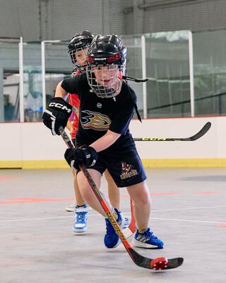 Two young players practice ball hockey indoors, wearing helmets and gloves, focused on teamwork and skill development.