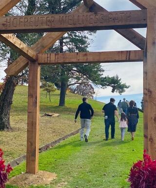 A family walks along a scenic path marked "Hofreuter Wellness Path," surrounded by greenery and trees under a cloudy sky.
