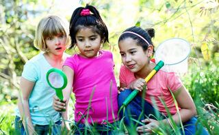Three children explore a grassy area, excitedly examining nature with a magnifying glass and bug-catching nets, immersed in discovery.