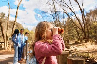 Three girls explore nature, with one examining something through a magnifying glass, while the others watch with curiosity and excitement.