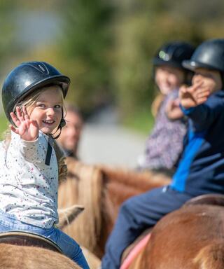 A joyful girl on a pony waves enthusiastically, while two other children, also on horseback, share the fun in a sunny outdoor setting.