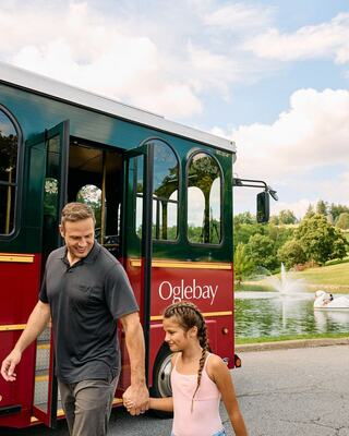 A man and a girl walk hand in hand from a trolley, enjoying a scenic park view with water features and lush greenery in the background.