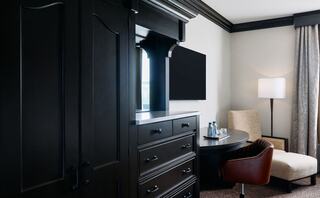 A modern hotel room features dark wood furniture, a small table with chairs, a TV, and soft lighting from a floor lamp.