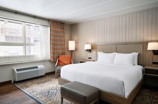 Bright, modern hotel room featuring a king-sized bed, large window with curtains, cozy seating, and warm wood accents.