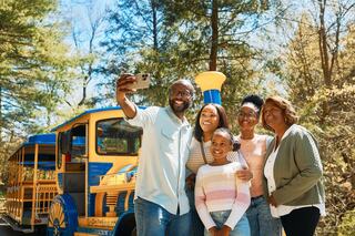 A young girl in a red dress joyfully leaps on a brightly colored bouncing surface, surrounded by greenery and wooden fencing.