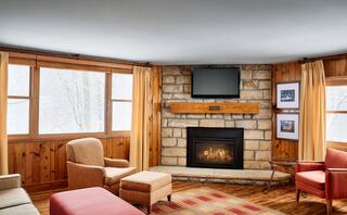 Cozy cabin interior with a wood-paneled warm ambiance, featuring a stone fireplace, comfortable seating, and large windows revealing a snowy landscape.