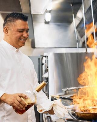 A chef joyfully adds liqueur to a sizzling pan, creating a dramatic flare of flames in a busy kitchen.