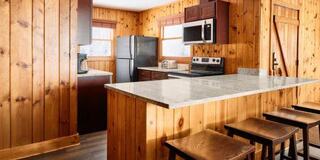 A cozy kitchen features wooden walls, dark cabinetry, stainless appliances, a granite countertop, and wooden stools, creating a warm atmosphere.