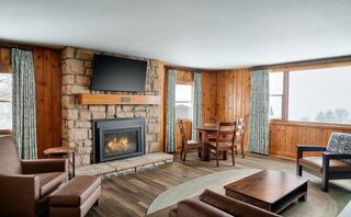 Cozy living area with a stone fireplace, comfortable seating, and a dining table, surrounded by large windows and rustic wood accents.