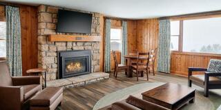 A cozy room with a stone fireplace, wooden walls, large windows, and comfortable seating, creating a warm and inviting atmosphere.