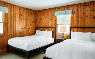 A cozy room featuring two comfortable beds, wooden paneling, and natural light from windows adorned with light blue curtains.