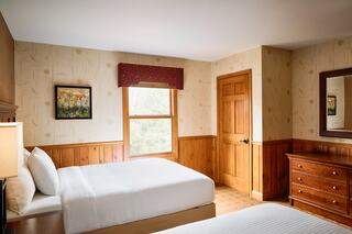 A cozy room features two beds, wooden decor, a window with a view, a dresser, and a painting on the wall, creating a warm atmosphere.