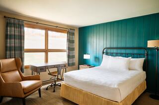 A cozy room featuring a comfortable bed, a desk with a chair, and large windows with plaid curtains, set against a teal accent wall.