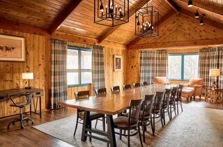 A warm, rustic meeting room features a long wooden table, comfortable seating, and large windows with patterned curtains, exuding a cozy atmosphere.