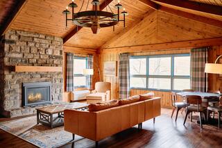 A cozy cabin interior featuring wooden walls, a stone fireplace, stylish furniture, and large windows for natural light.