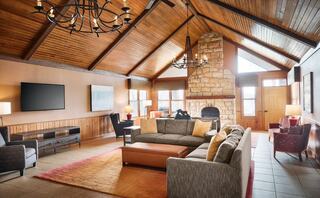 A spacious living room features high wooden ceilings, a stone fireplace, large windows, cozy seating, and warm, inviting decor.