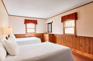 Cozy room featuring two beds, wooden paneling, warm lighting, and bright windows adorned with red valances.