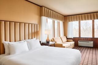 A cozy hotel room featuring a spacious bed, a comfy sofa, ample natural light from large windows, and warm, inviting decor.