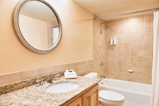 A modern bathroom featuring a granite countertop, circular mirror, shower tub combo, beige tiled walls, and neatly arranged towels.