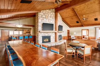 A spacious living area features a large wooden table, cozy seating, two fireplaces, and large windows, all under a high, wooden ceiling.