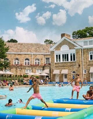 Kids play on inflatable mats in a sunny pool, surrounded by joyful swimmers and a beautiful stone building in the background.