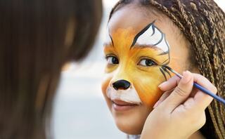 A girl smiles with her face painted like a lion as someone adds the finishing touches to her make-up, showcasing bright colors and artistry.