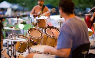 A drummer plays a set in a lively outdoor setting, with a musician on guitar in the background, creating a vibrant atmosphere.