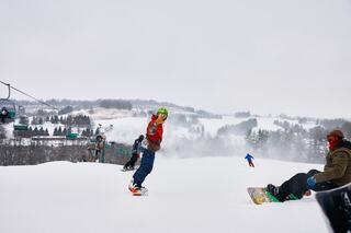 A young snowboarder poses excitedly on a snowy slope while others relax nearby. Ski lifts and a winter landscape complete the scene.
