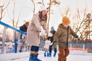 A woman encourages a young boy on ice skates, while others enjoy skating nearby in a winter setting. Laughter and joy fill the scene.