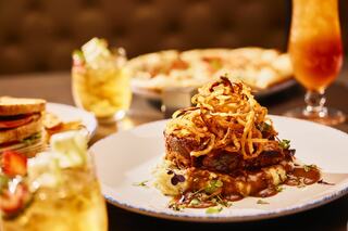 A gourmet steak topped with crispy onions, served on a bed of creamy mashed potatoes and accompanied by drinks and a sandwich in the background.