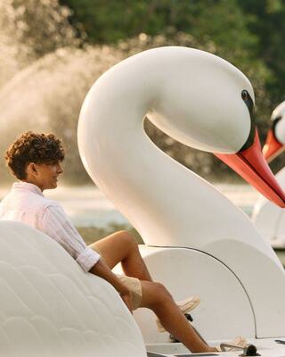 A young man relaxes on a large swan-shaped paddle boat, with fountains and greenery in the background, enjoying a sunny day.
