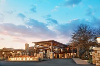 A modern lodge with a welcoming entrance, stone accents, and a backdrop of soft clouds during sunset, surrounded by trees.