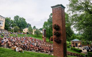 A large outdoor amphitheater filled with an audience enjoys a performance, surrounded by greenery and charming brick structures.