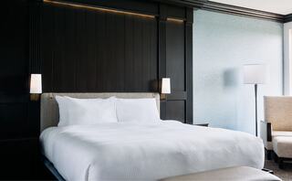 A modern bedroom features a plush bed with crisp white linens, dark paneled walls, stylish lamps, and minimalist furniture for a cozy atmosphere.