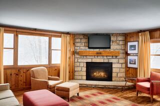 Cozy room with stone fireplace, modern TV, and comfortable seating. Large windows provide a view of the outdoors, enhancing the warm ambiance.