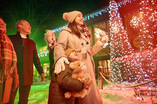 A group of people, including a woman and a child holding a teddy bear, enjoy festive lights and snow, creating a cheerful holiday atmosphere.
