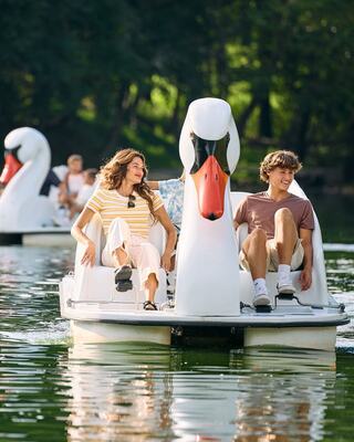 A couple enjoys a leisurely ride on a swan pedal boat, surrounded by lush greenery and other boats on the water.