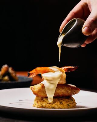 A hand pours sauce over a tower of shrimp atop a crispy base, creating an enticing dish garnished elegantly on a plate.