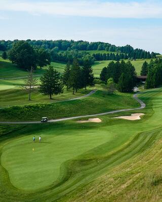 A lush golf course sprawls across rolling hills, featuring well-manicured greens, trees, and a winding path, with golfers in the distance.