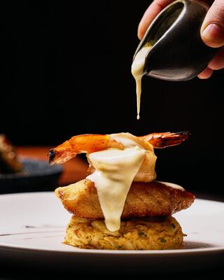 A hand pours sauce over a layered dish featuring shrimp atop a crispy base, showcasing a delicious and elegant presentation.