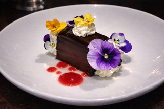 A rich chocolate dessert garnished with vibrant edible flowers and a drizzle of raspberry sauce, elegantly arranged on a white plate.