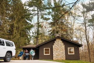 A group of three people approaches a cozy cabin surrounded by tall trees, with a stylish SUV parked nearby. Nature and adventure await.