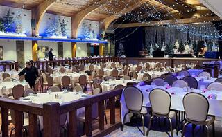 A spacious banquet hall is elegantly decorated with tables set for guests, featuring twinkling lights and festive holiday decor in the background.